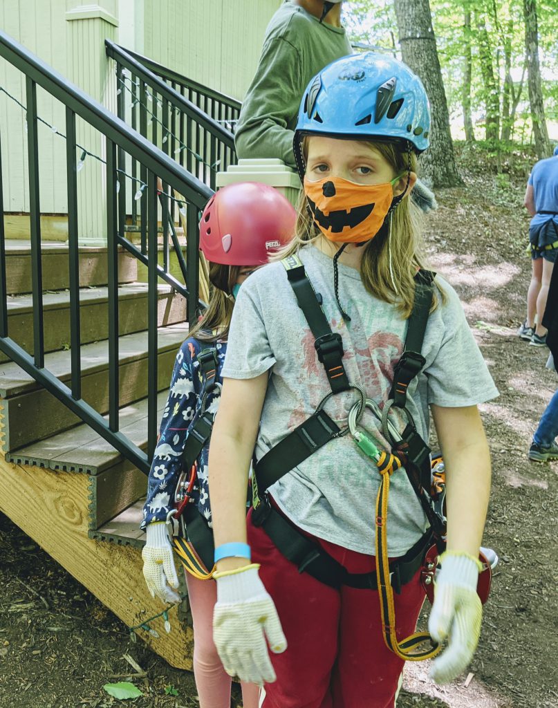 Silas, looking mildly distressed, wearing high ropes gear and a mask with a jack-o-lantern mask. Petra, wearing her gear, peeking out from behind him.