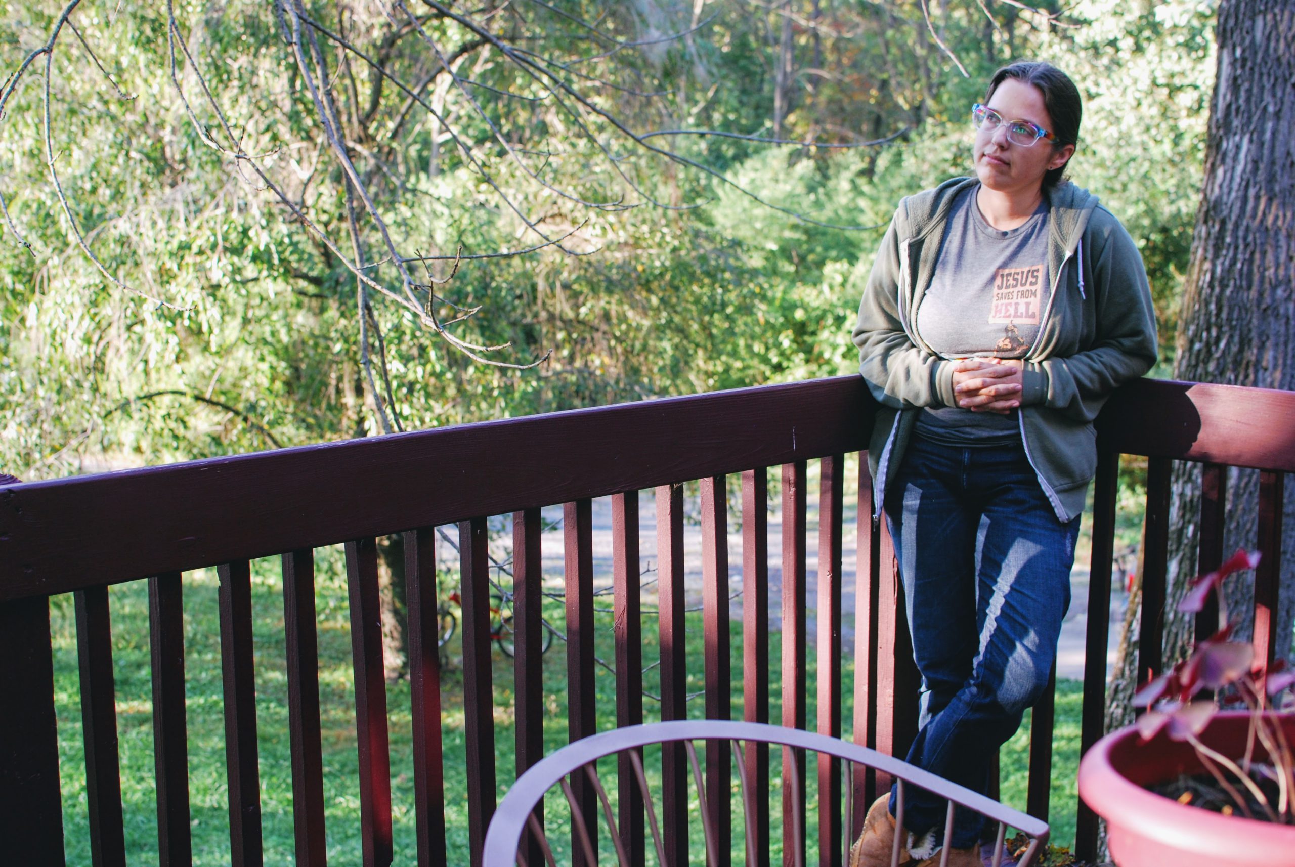 Aili on her porch, wearing jeans, a gray t-shirt, and a green hoodie.