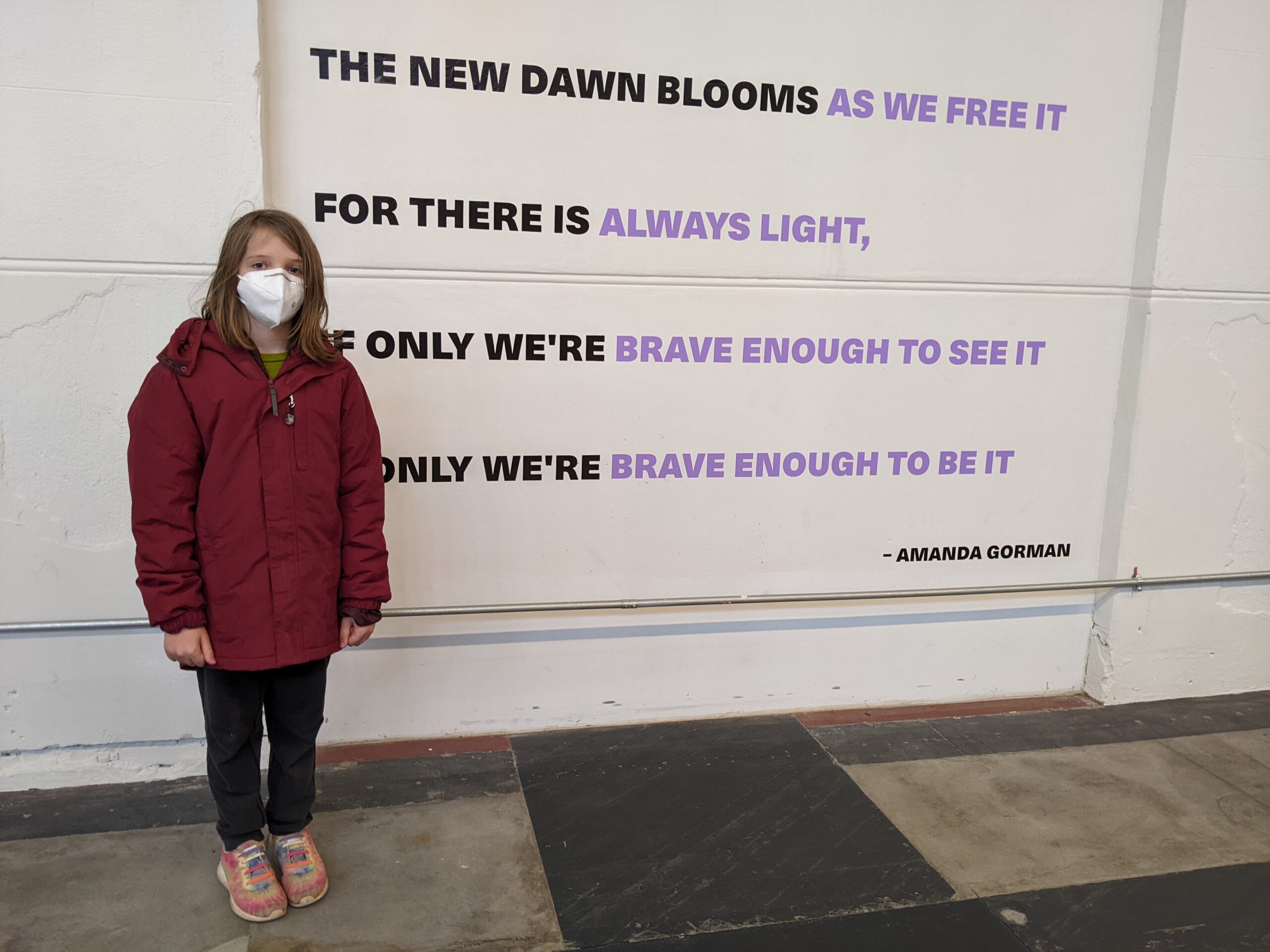 Silas standing in front of a wall that has a quotation on it: "The new dawn blooms as we free it / For there is always light, / If only we're brave enough to see it / If only we're brave enough to be it. -- Amanda Gorman"