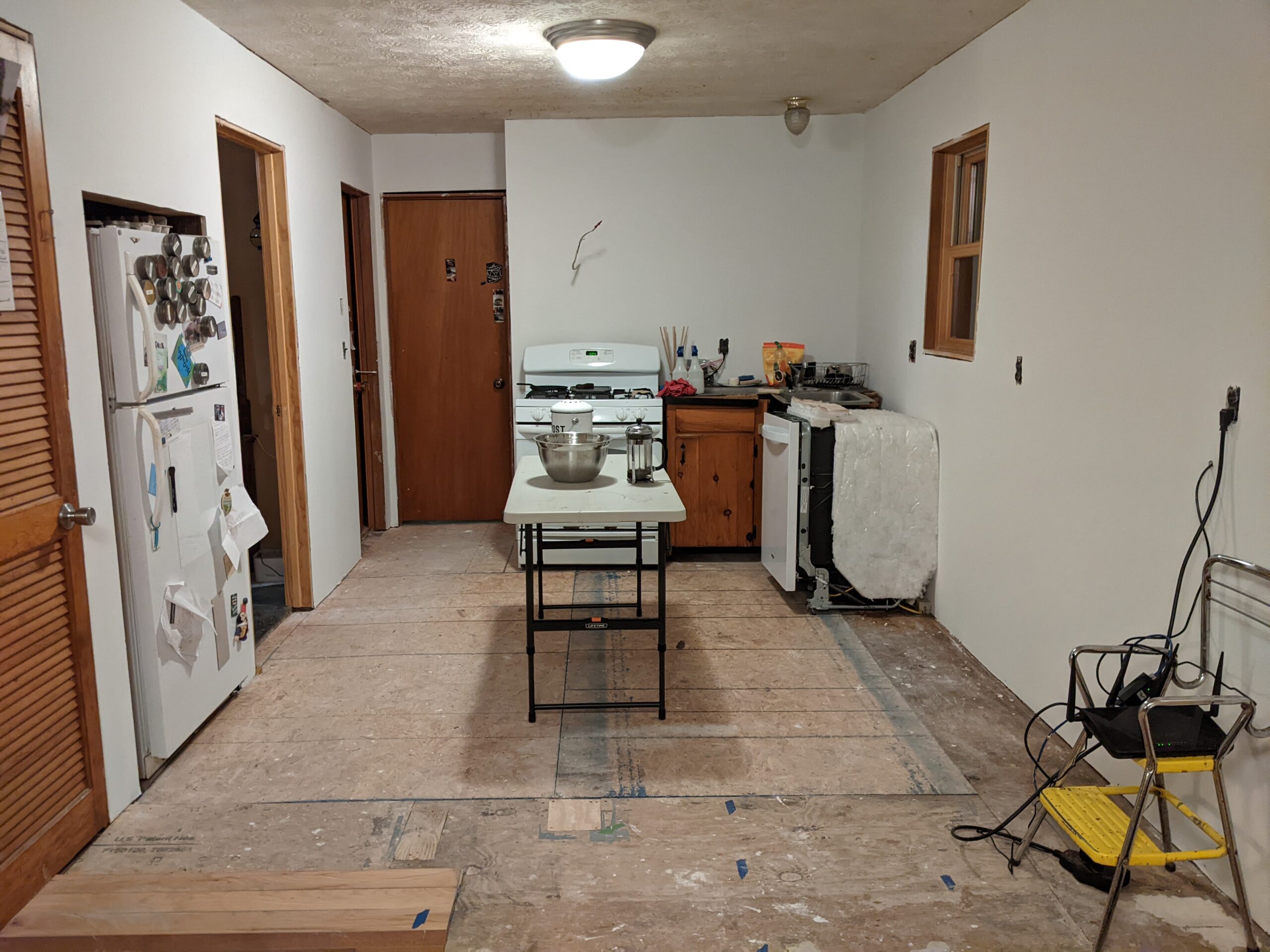 Kitchen with drywall hung and primed, and ...not much else in it. A stove, a dishwasher, a weird sink, a fridge, and a folding table.