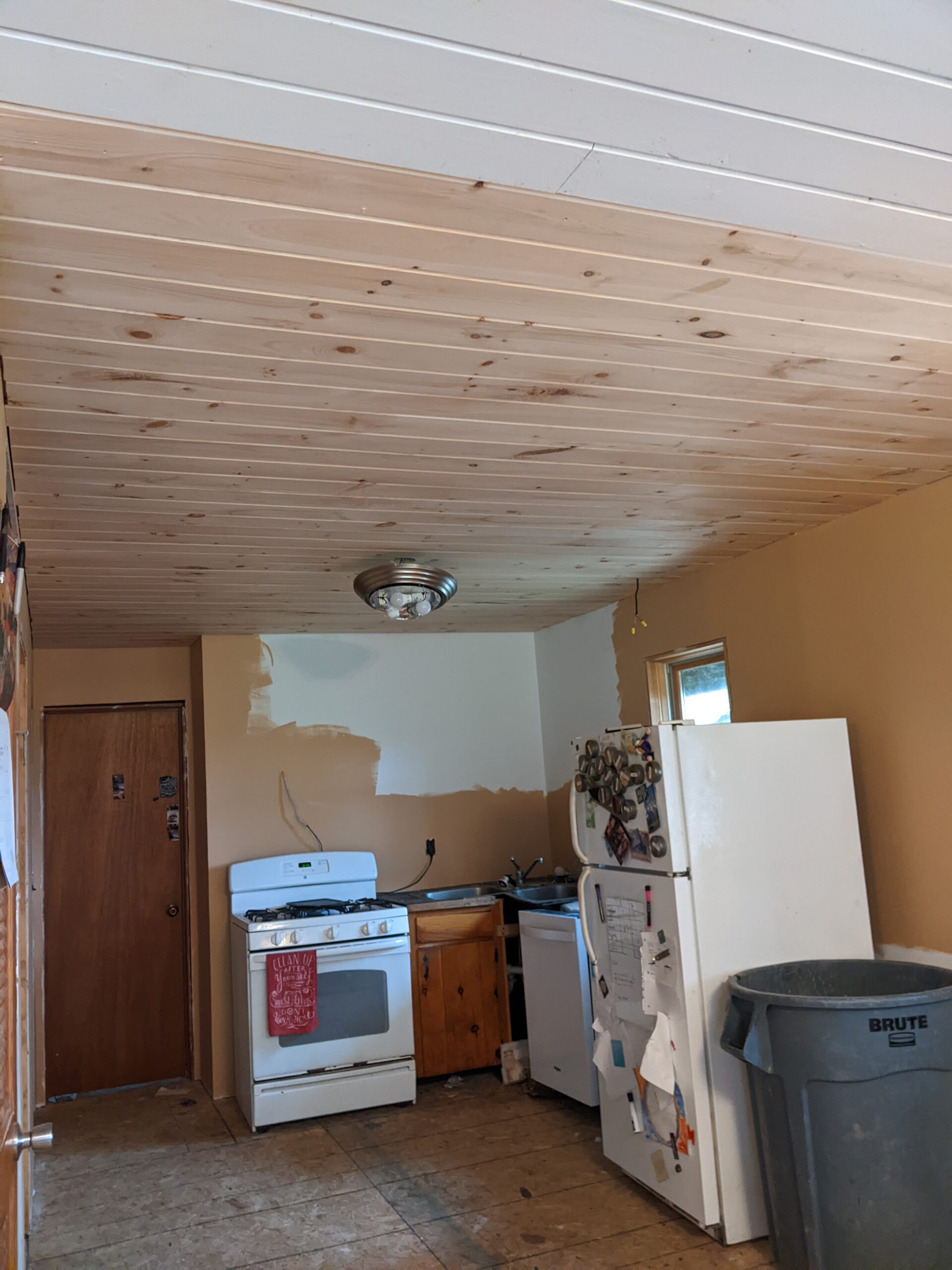 Photo of kitchen in progress, prominently showing the transition from the white painted pine ceiling in the living room to the unpainted new pine ceiling in the kitchen.