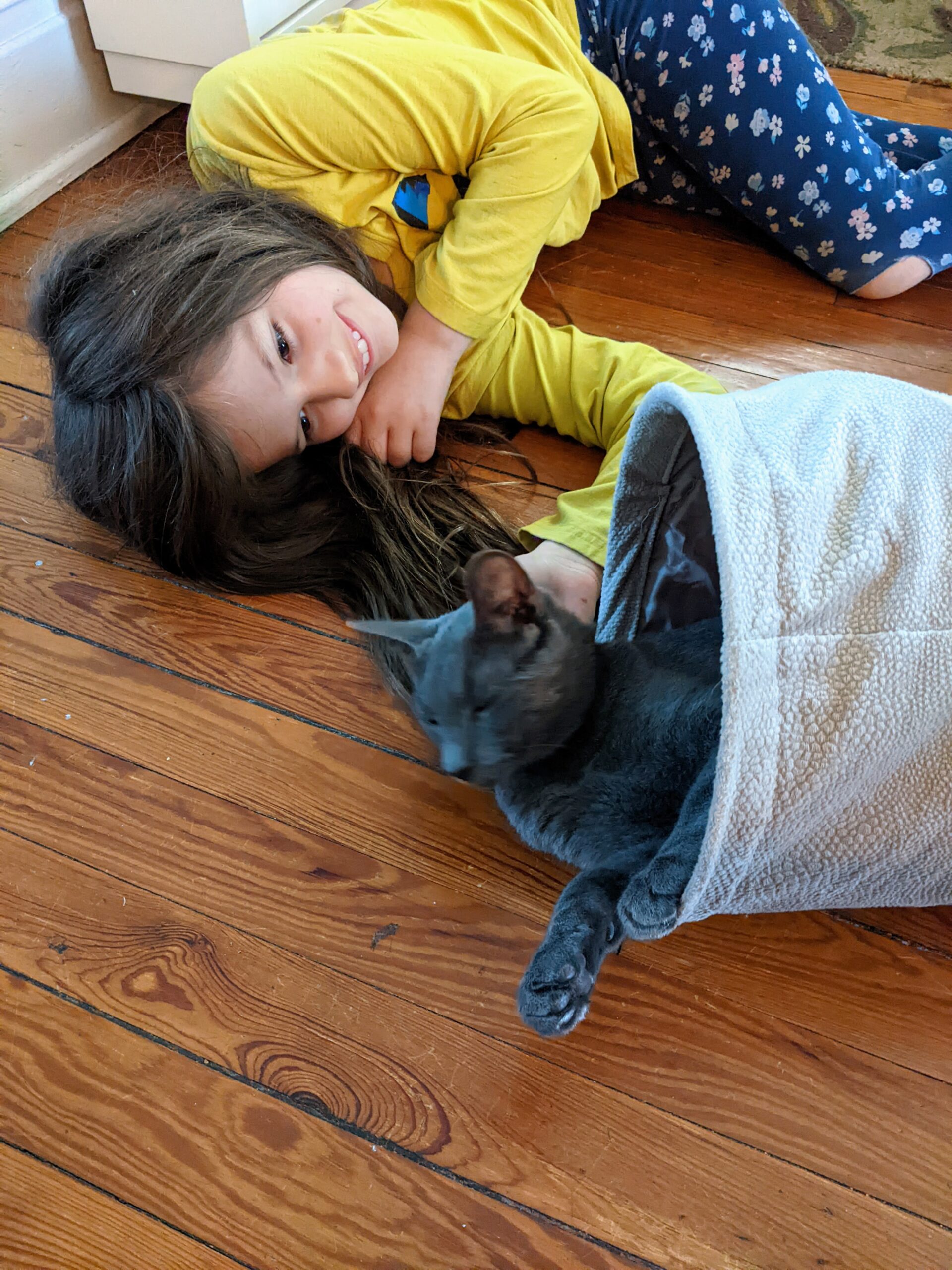 Petra laying on the floor, smiling at a gray cat who is in a tunnel.
