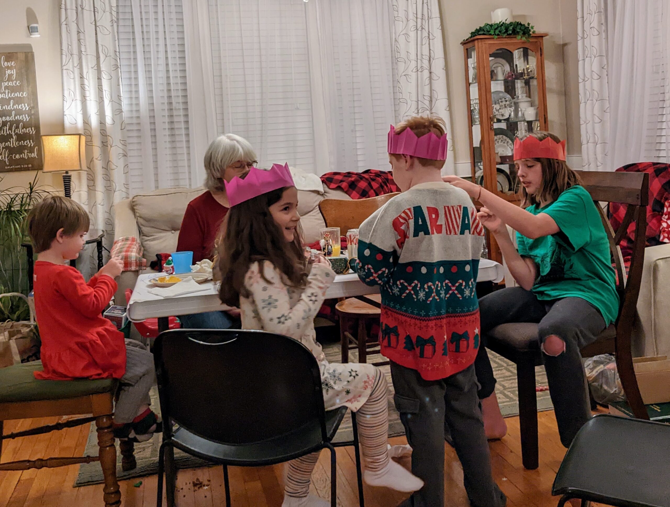 Elora, JJ, and Silas at the kids' table, wearing paper crowns, with Verna sitting in the background.