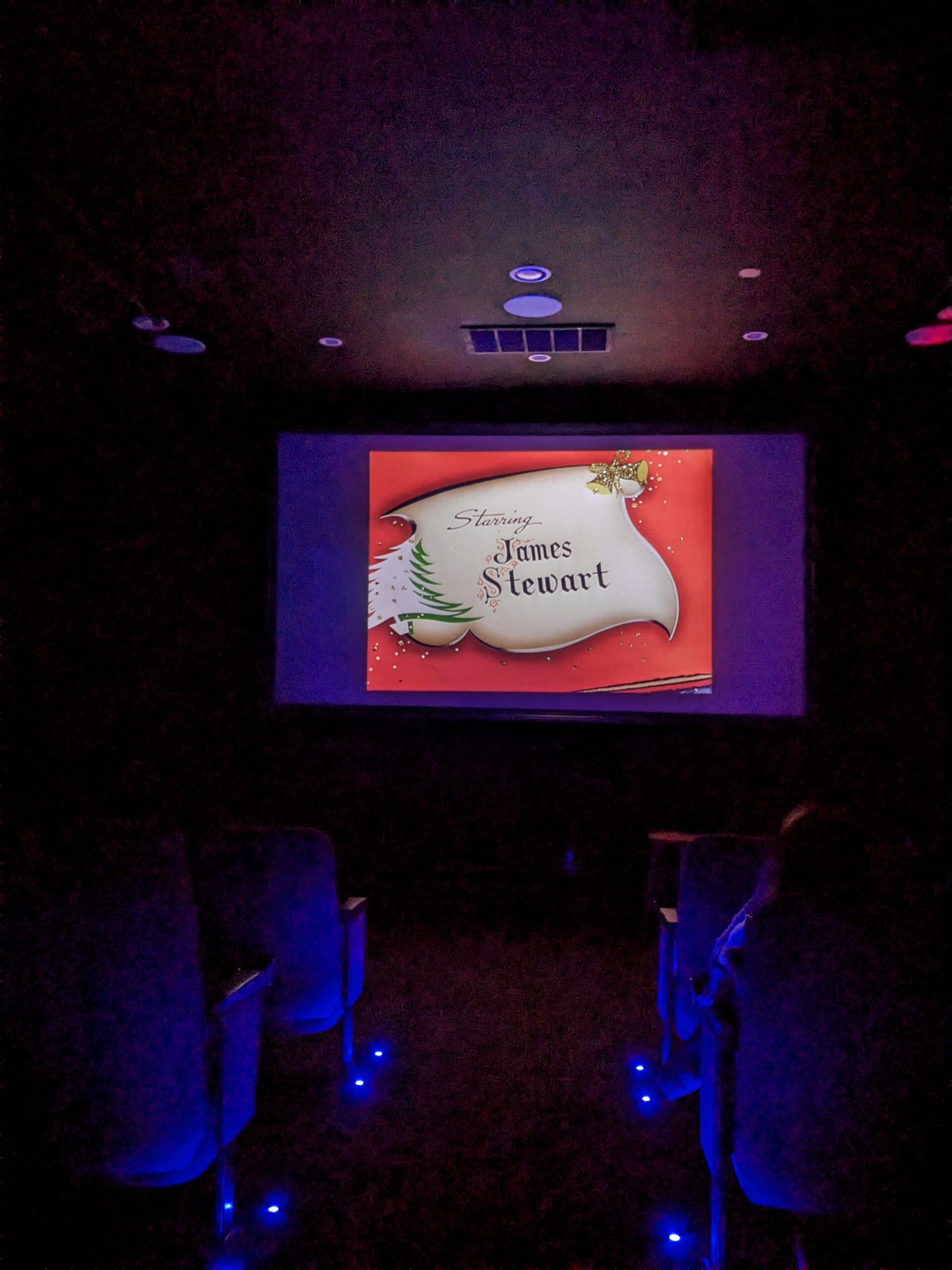 Dark theater. On the screen is one of the title cards from It's a Wonderful Life, which says, "Starring James Stewart"