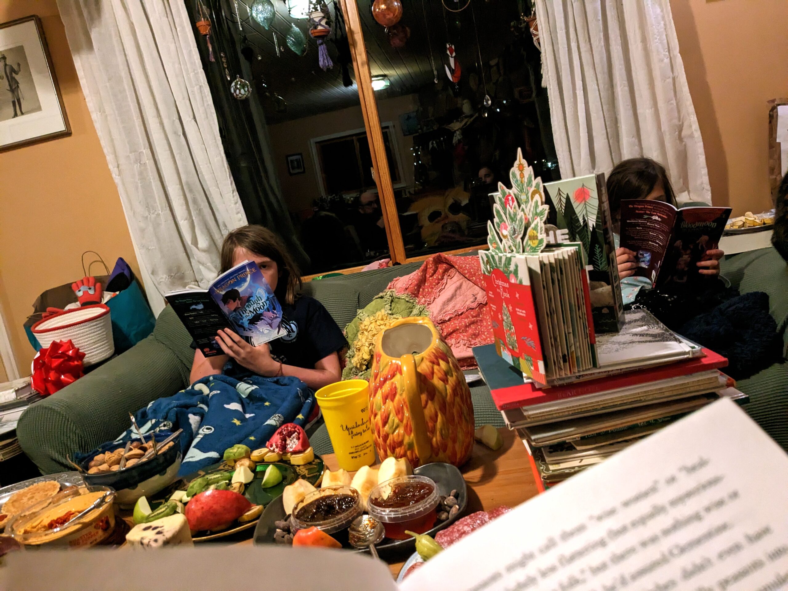 Silas and Petra, on the couch, reading, with food on the table nearby.
