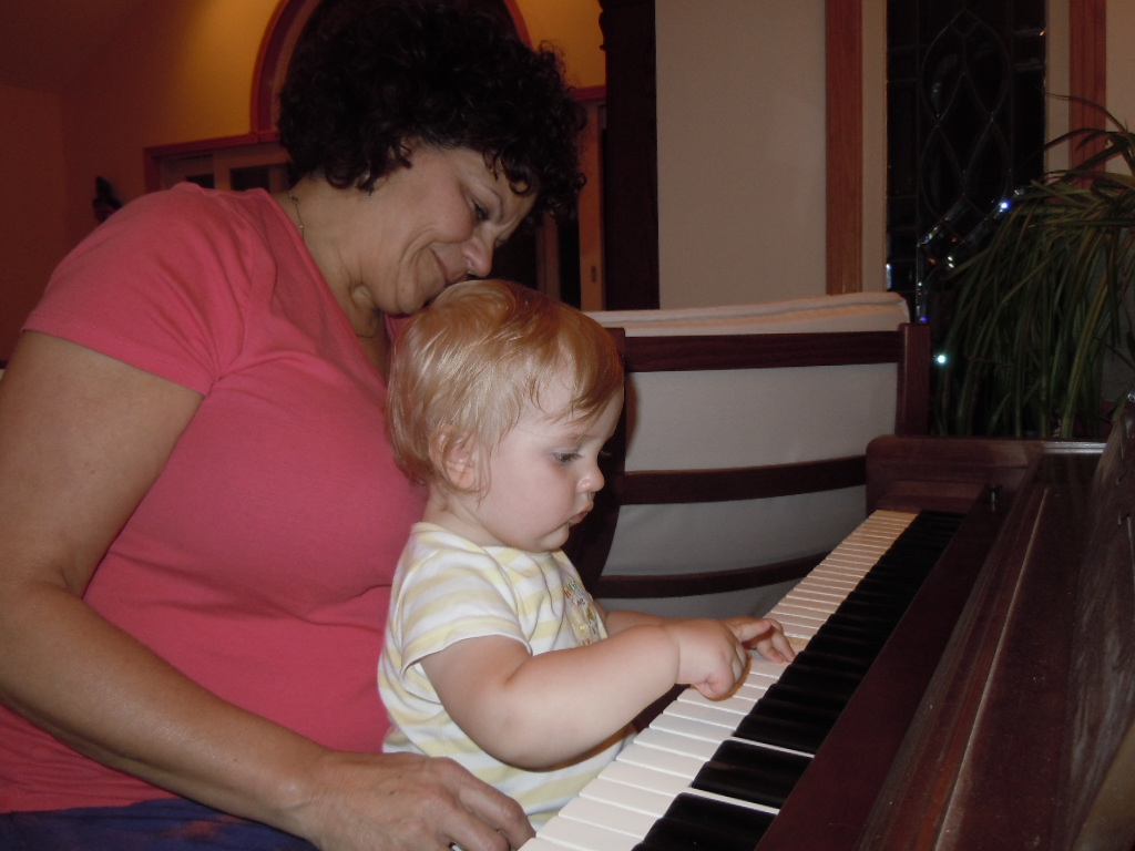 Toddler Silas is sitting on the lap of a lady in her late 50s. He is playing a piano.