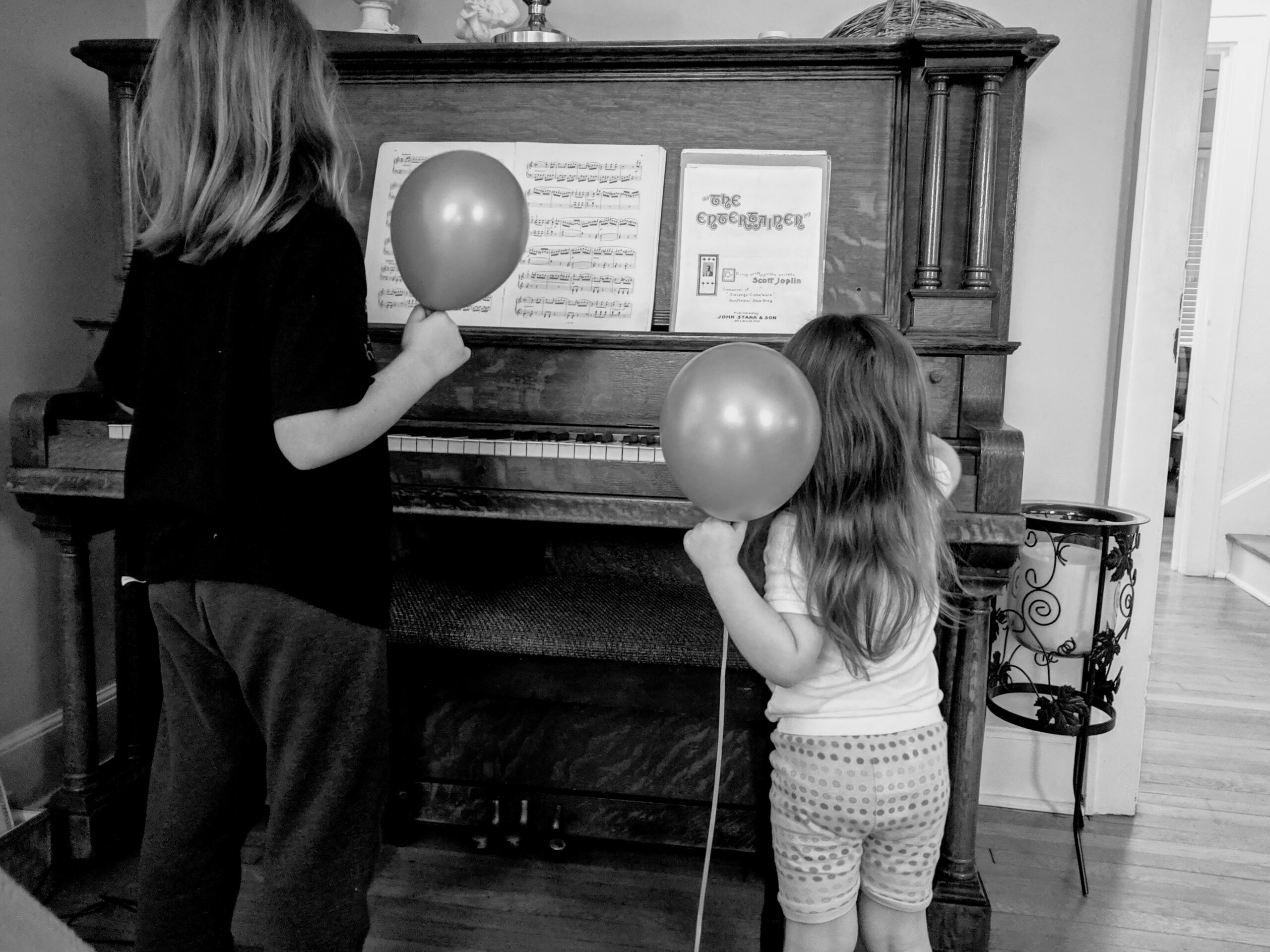 Black and white image of two children playing an upright grand. They are both holding balloons.