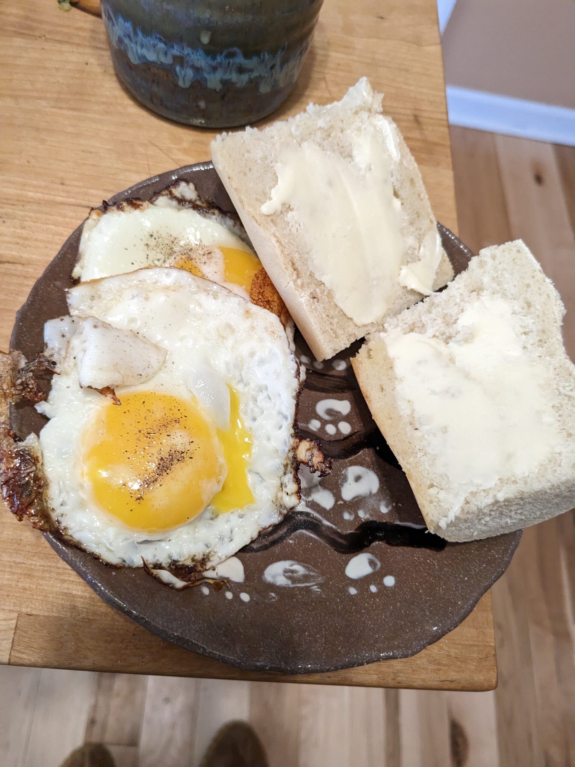 Two fried eggs and buttered slices of baguette on a brown pottery plate.